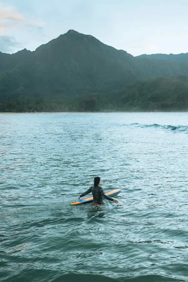 A surfer wading out with a longboard towards green mountains on the shore.