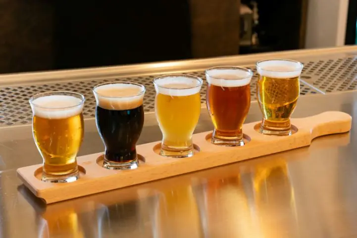 A tasting flight of five craft beers, from pale to dark, on a wooden paddle at a bar.